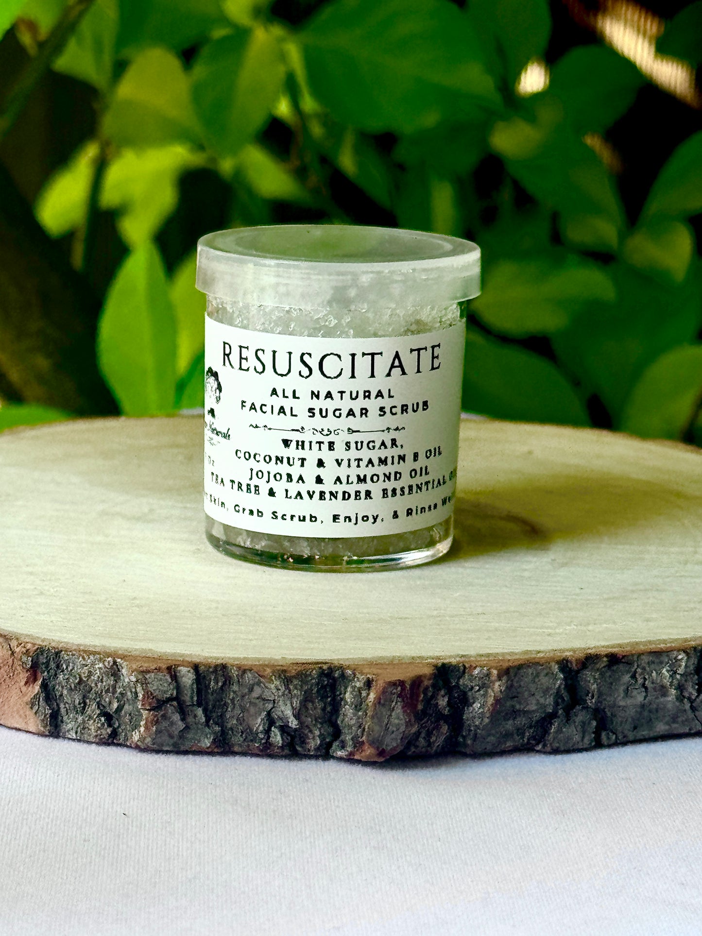 A small clear jar with a clear lid white label with black writing filled with Resuscitate All Natural Exfoliating Sugar Scrub on a wooden surface with green leaves in the background