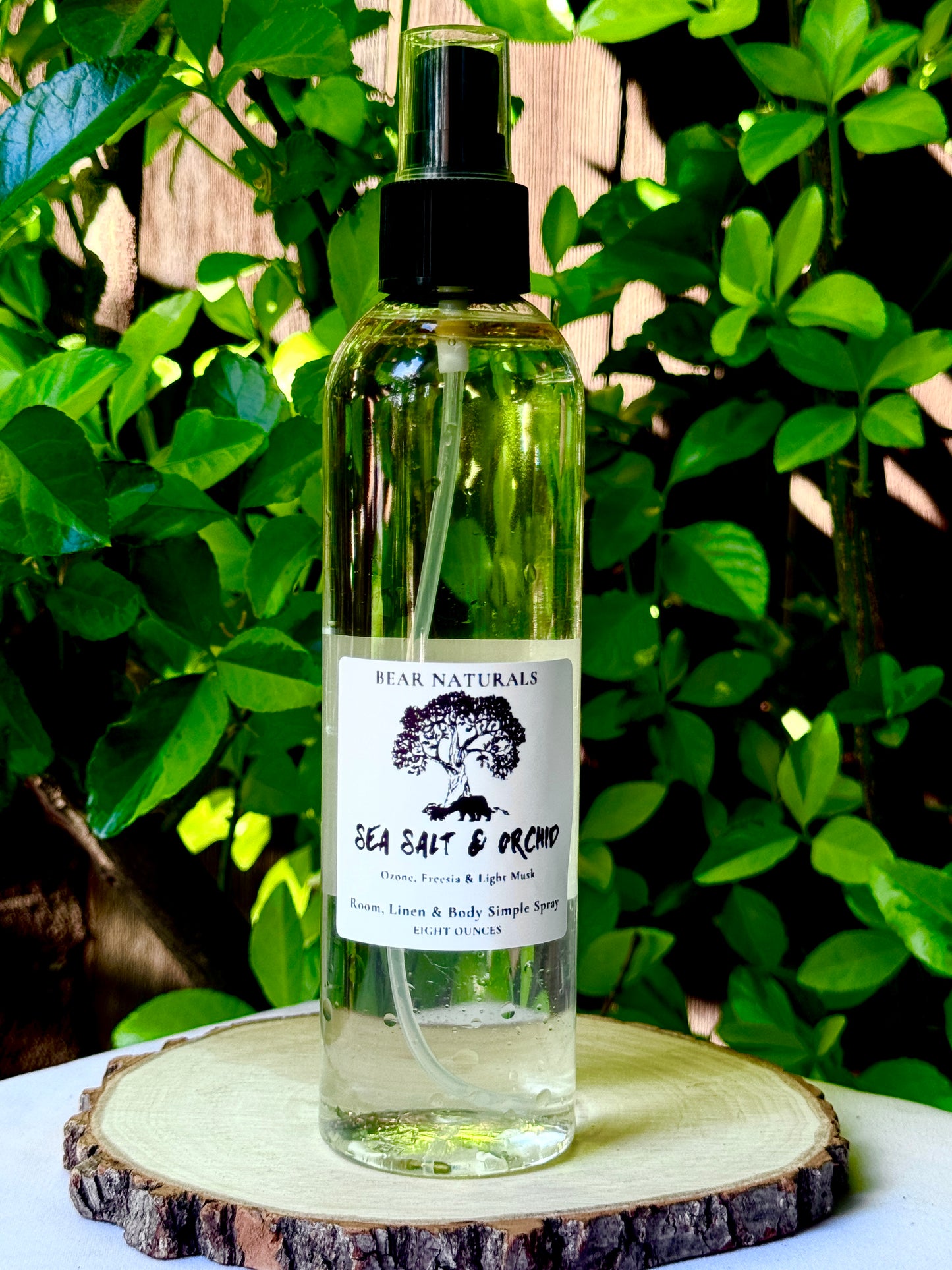 A clear bottle with a black fingertip style spray nozzle featuring a white label with black writing on a wooden surface with green leaves in the background.