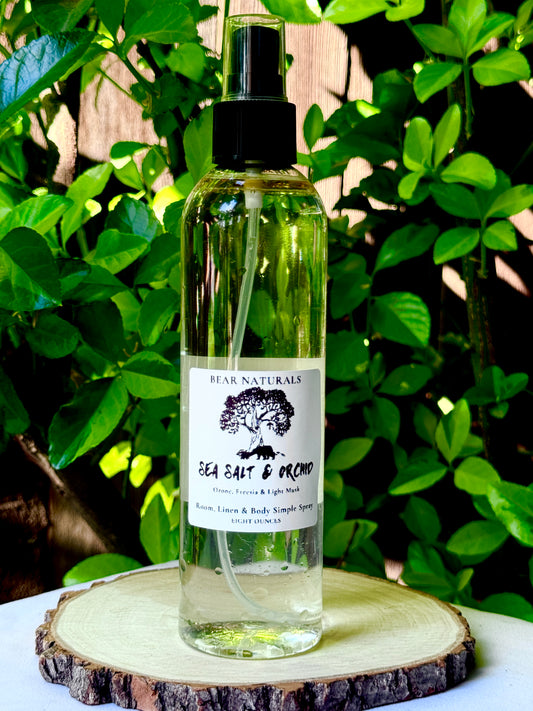 A clear bottle with a black fingertip style spray nozzle featuring a white label with black writing on a wooden surface with green leaves in the background.