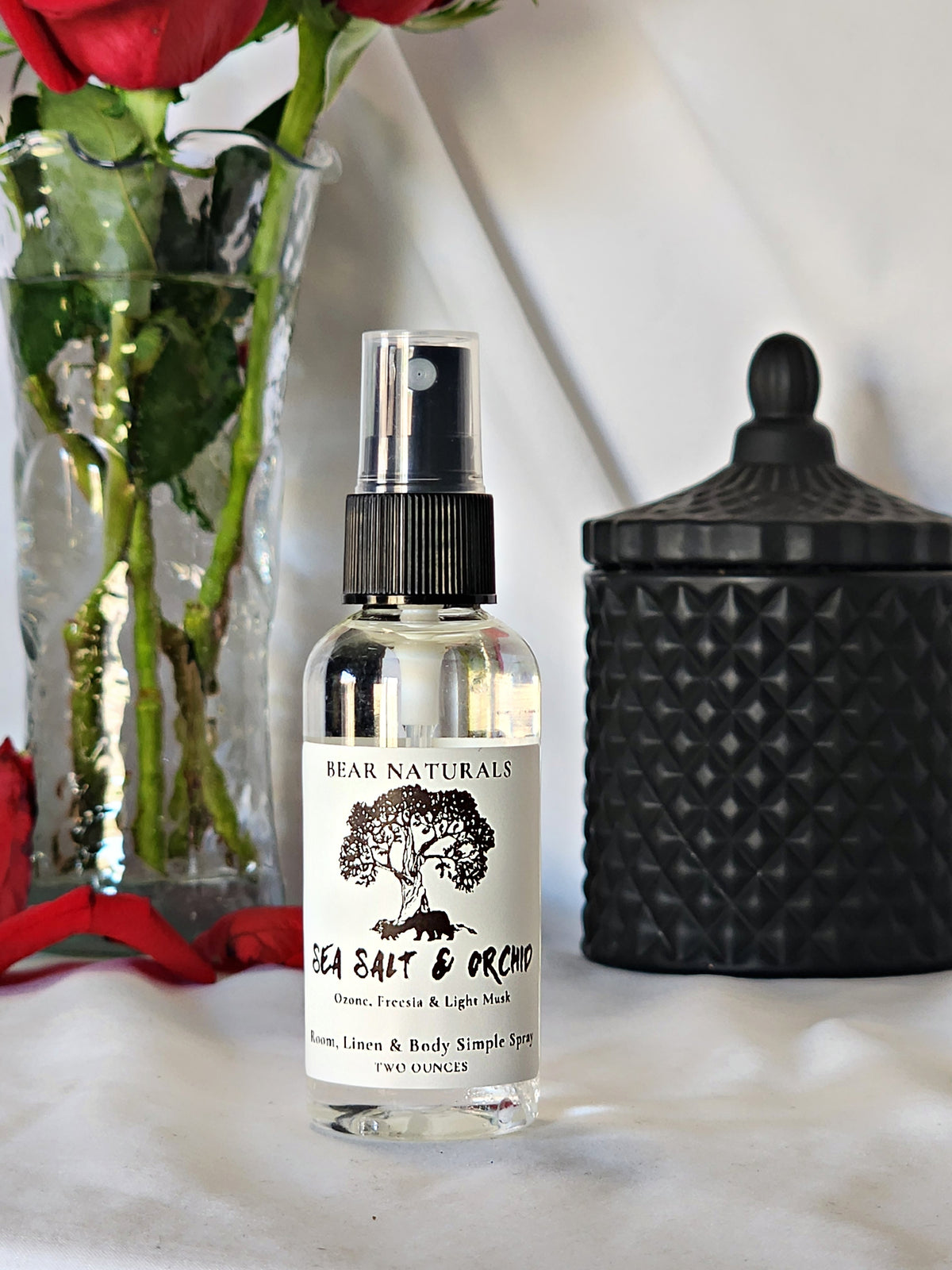 A clear bottle with a black fingertip style spray nozzle featuring a white label with black writing with a black candle jar and red roses in a vase in the background.