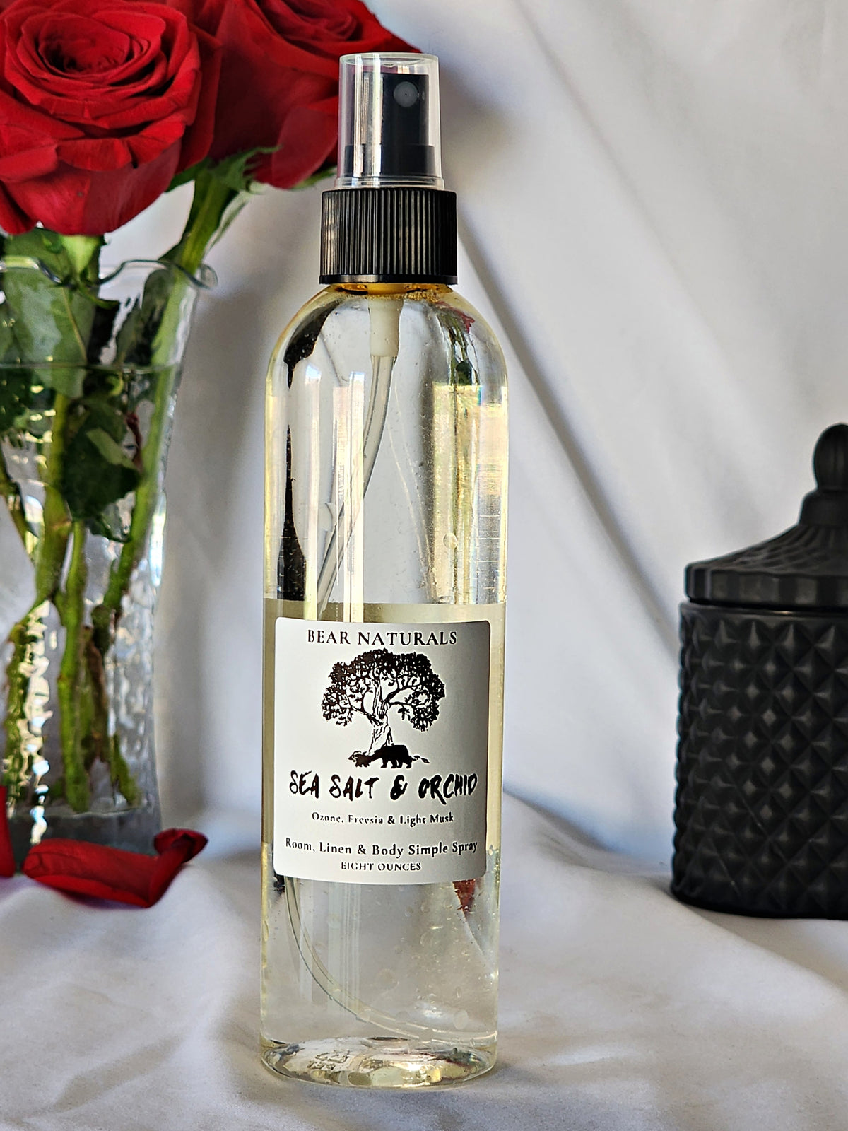 A clear bottle with a black fingertip style spray nozzle featuring a white label with black writing with a black candle jar and red roses in a vase in the background.