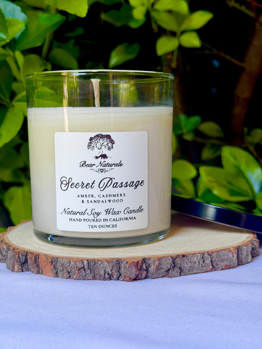 A white ten ounce candle in a clear glass jar with a black metal lid and white label with black writing sitting on a wooden surface. The candle is called "Secret Passage".