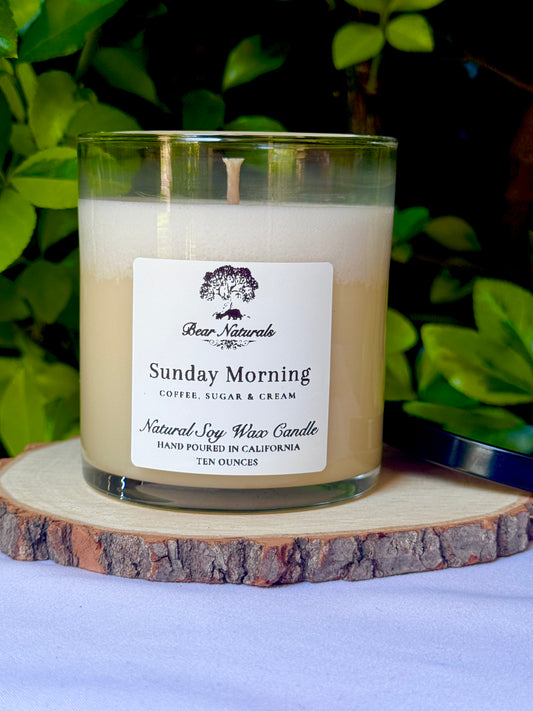 A white ten ounce candle in a clear glass jar with a black metal lid and white label with black writing sitting on a wooden surface. The candle is called "Sunday Morning".