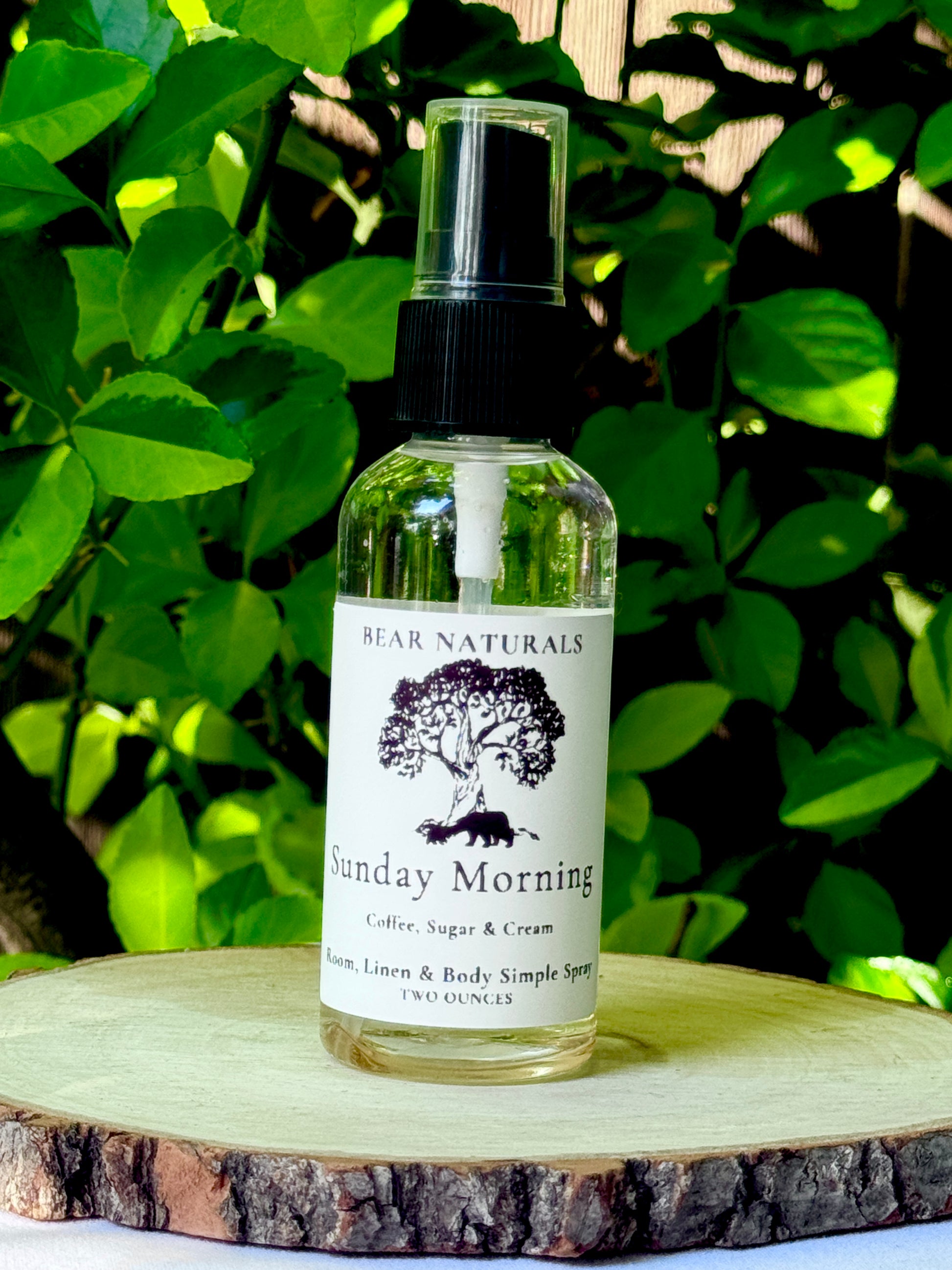A clear bottle with a black fingertip style spray nozzle featuring a white label with black writing on a wooden surface with green leaves in the background.
