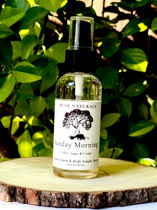 A clear two ounce finger-tip spray bottle with black and white label on a wooden surface with green leaves in the background. The bottle contains the scent "Sunday Morning".