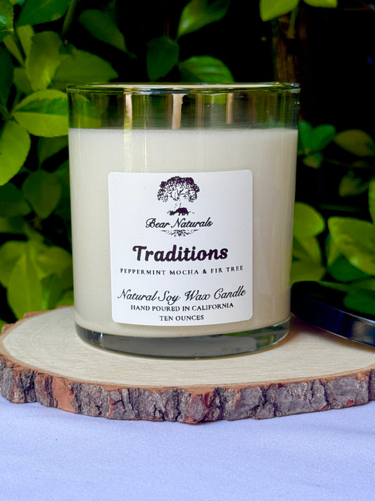 A white ten ounce candle in a clear glass jar with a black metal lid and white label with black writing sitting on a wooden surface. The candle is called "Traditions".