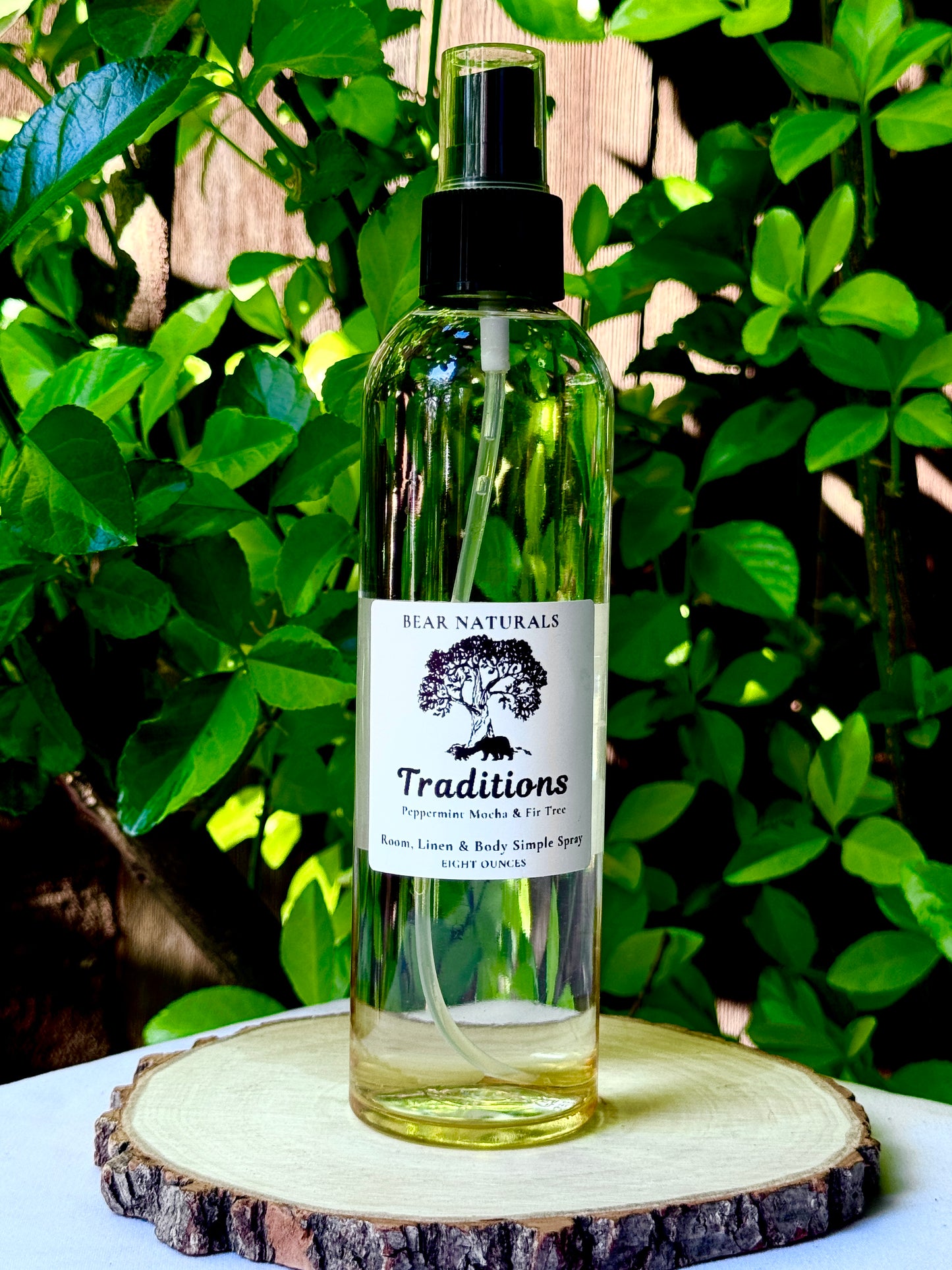A clear bottle with a black fingertip style spray nozzle featuring a white label with black writing on a wooden surface with green leaves in the background.