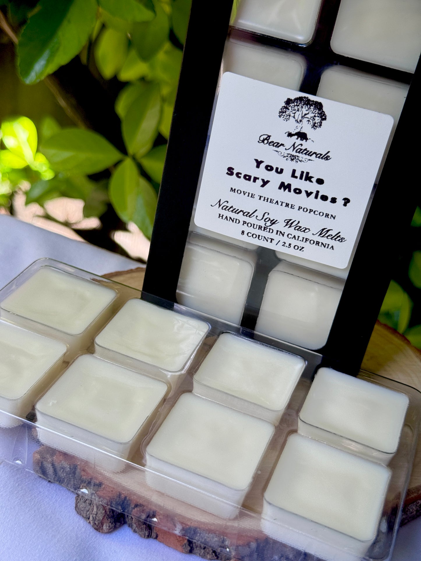 A black box with a clear plastic tray housing eight white wax melts featuring a white label with black writing on a wooden surface with green leaves in the background
