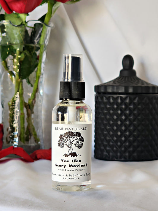 A clear bottle with a black fingertip style spray nozzle featuring a white label with black writing with a black candle jar and red roses in a vase in the background.