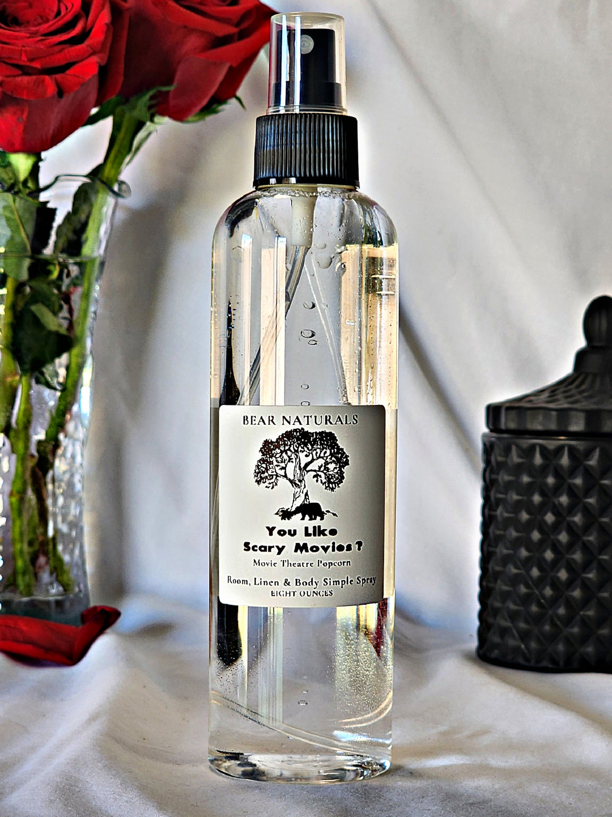 A clear bottle with a black fingertip style spray nozzle featuring a white label with black writing with a black candle jar and red roses in a vase in the background.